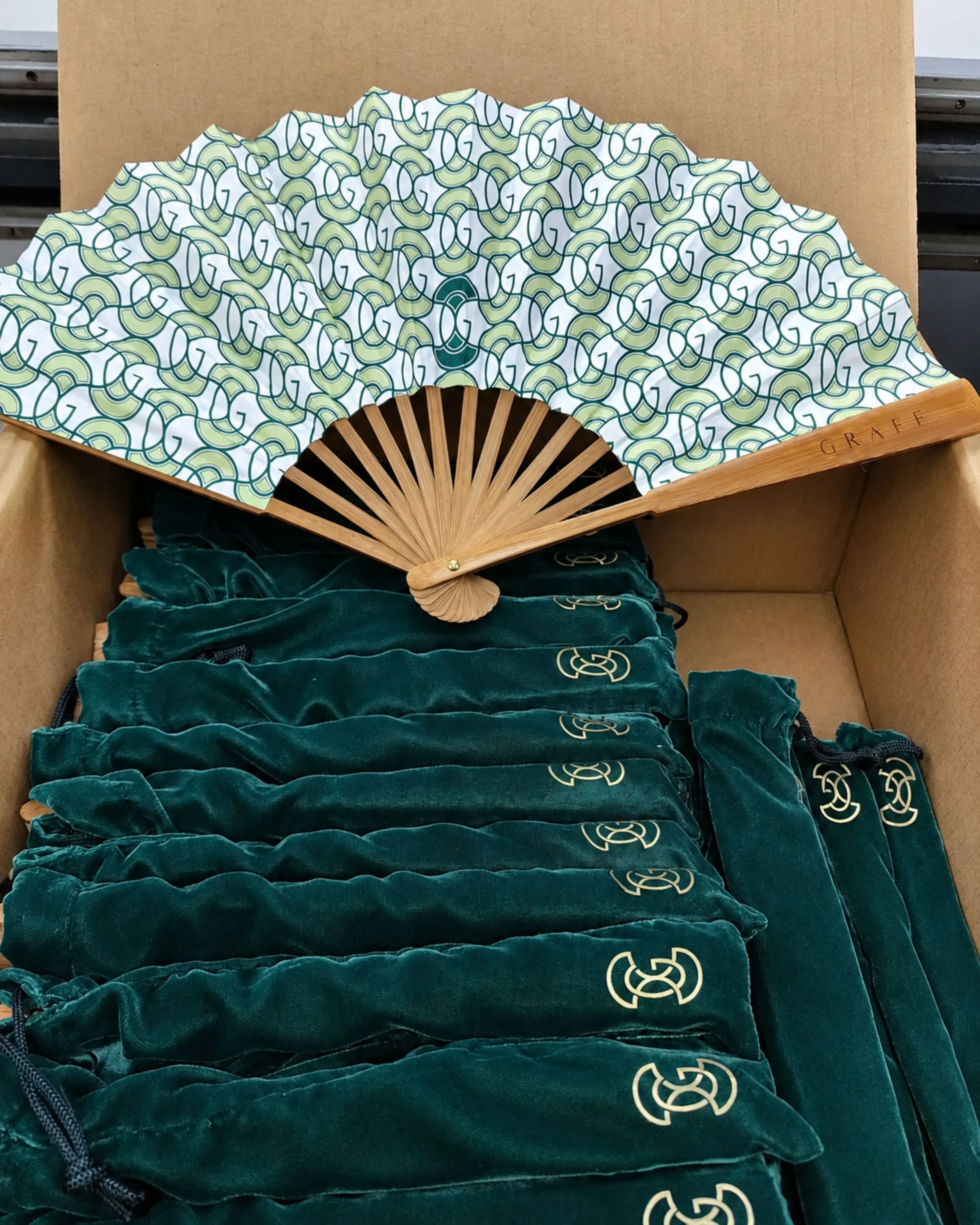 Open box of bamboo fans with rows of green velvet G-monogrammed drawstring pouches ready for delivery